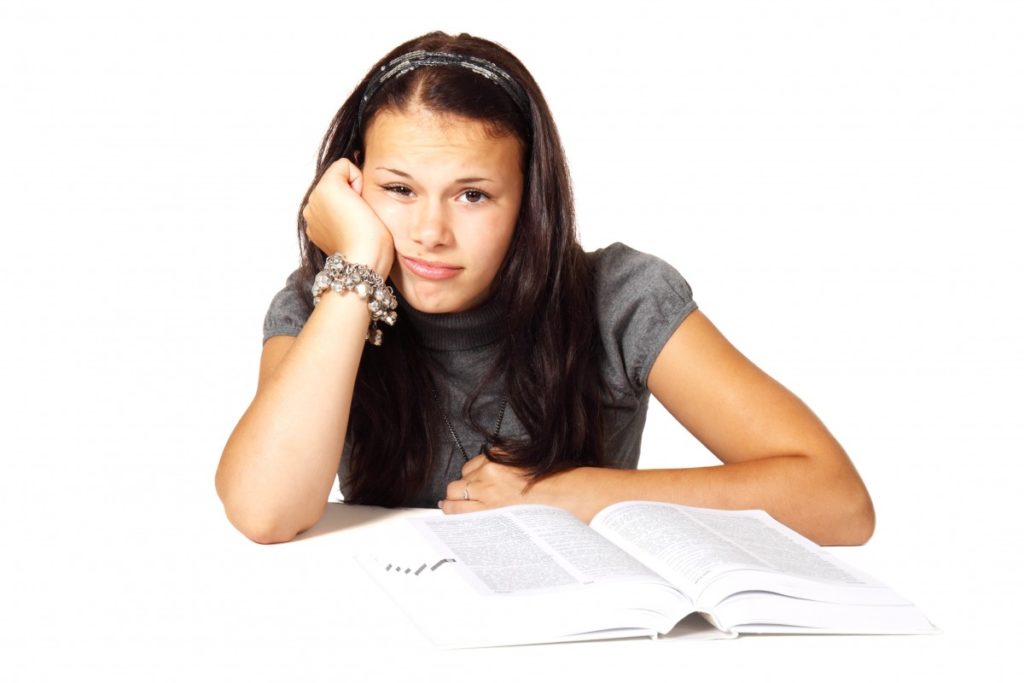 Annoyed-looking girl stares out at us (front-view), cheek resting against her hand while an open book lies on table in front of her.